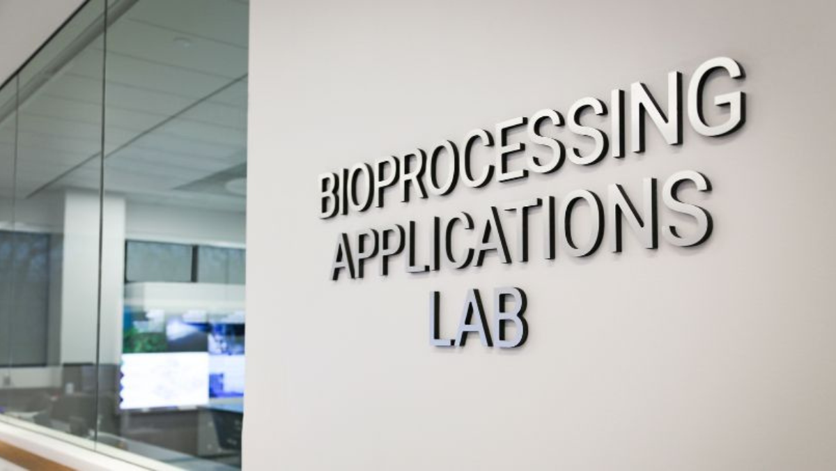 Bioprocessing Applications Lab sign on a modern facility wall viewed through adjacent glass window.